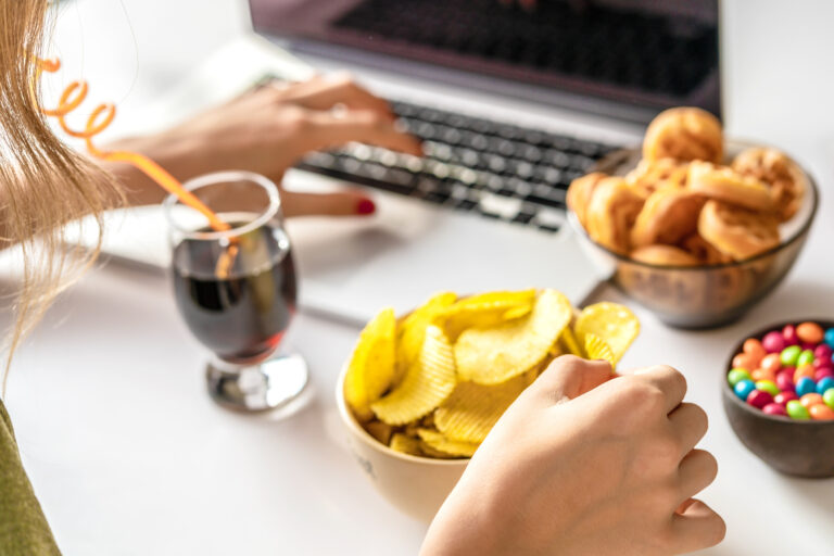 Jente som ser jobber på data på samme gang som hun drikker cola og spiser potetgull og gotterier